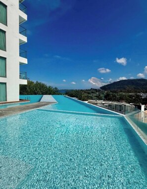A swimming pool with clear blue water, surrounded by a white building with balconies, set against a backdrop of mountains and a clear sky. - Fully Seaview 2BR Apartment, Walk to Kamala Beach (Kamala)