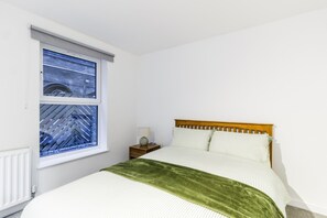 Ein Schlafzimmer mit einem Bett, einem Nachttisch, einer Lampe, einem Fenster mit Blick auf ein Gebäude und einer Heizung.