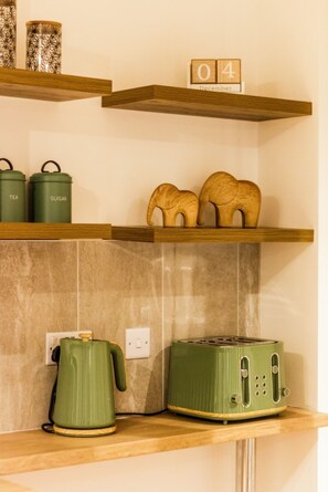 Eine Küchenarbeitsplatte mit einem grünen Wasserkocher, einem grünen Toaster und zwei grünen Behältern mit den Aufschriften 'TEE' und 'ZUCKER'. Darüber befindet sich ein Holzregal mit einer Holznummer '04' und einem Kalender, der 'Dezember' anzeigt.