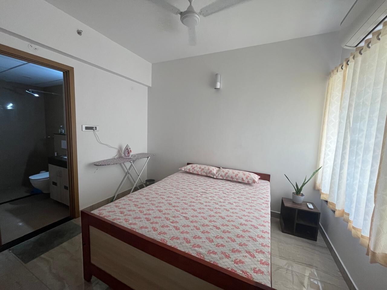 A bedroom with a bed, a ceiling fan, a small table with a plant, and a window with curtains.