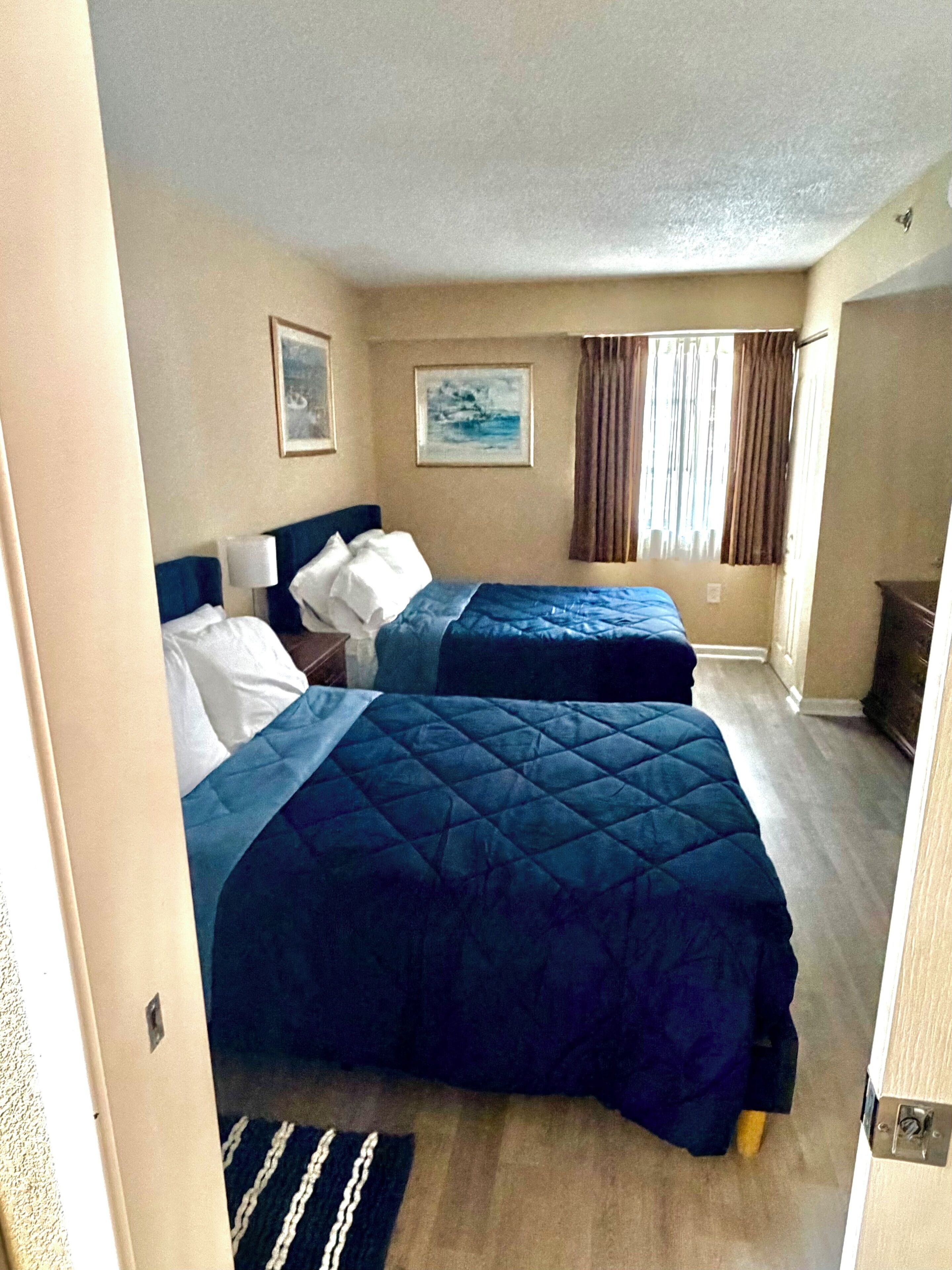 A hotel room with two beds, a nightstand, a lamp, a painting, and a window with curtains.
