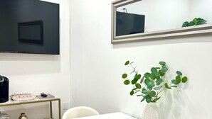 Una zona de menjador moderna amb taula blanca, quatre caires, un vas amb plantes verdes, televisió penjada a la paret i una petita estanteria amb un aparell de ràdio i objectes decoratius.