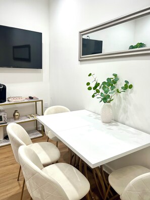Ein stilvoller Essbereich mit einem weißen Tisch, vier Stühlen, einer Vase mit Grünpflanzen, einem Fernseher an der Wand und einer kleinen Ablage mit Radio und Dekoration.
