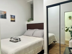 Ein ordentlich gemachtes Bett mit einem gefalteten Handtuch und einem kleinen Stapel Kleidung, eine Zimmerpflanze und ein an der Wand befestigter Spiegel.