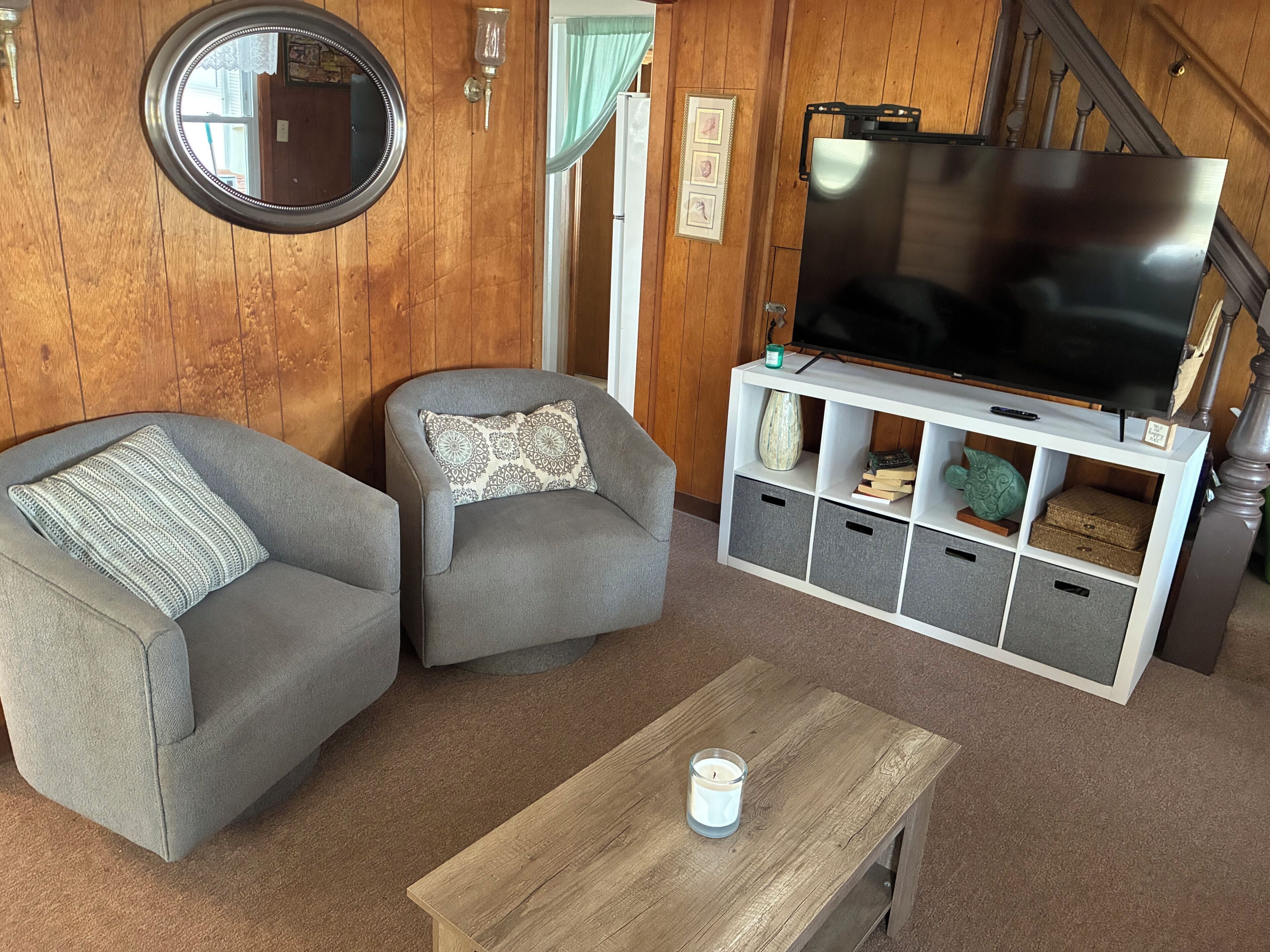 A living room with two grey armchairs, a wooden coffee table with a candle, a TV on a white stand, and wooden paneling on the walls.