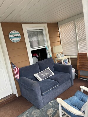 Ein blaues Sofa mit einem 'Relax'-Kissen, ein kleiner Tisch mit einer Lampe und eine Uhr an der Wand.