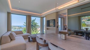 Ein modernes Wohnzimmer mit Sofa, Esstisch und Stühlen, großen Fenstern mit Vorhängen, einem Flachbildfernseher und Meerblick.