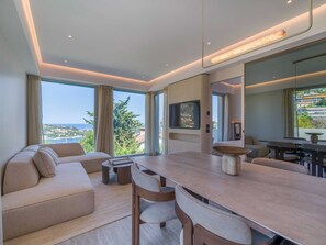 Ein modernes Wohnzimmer mit Sofa, Esstisch und Stühlen, großen Fenstern mit Vorhängen, einem Flachbildfernseher und Meerblick.