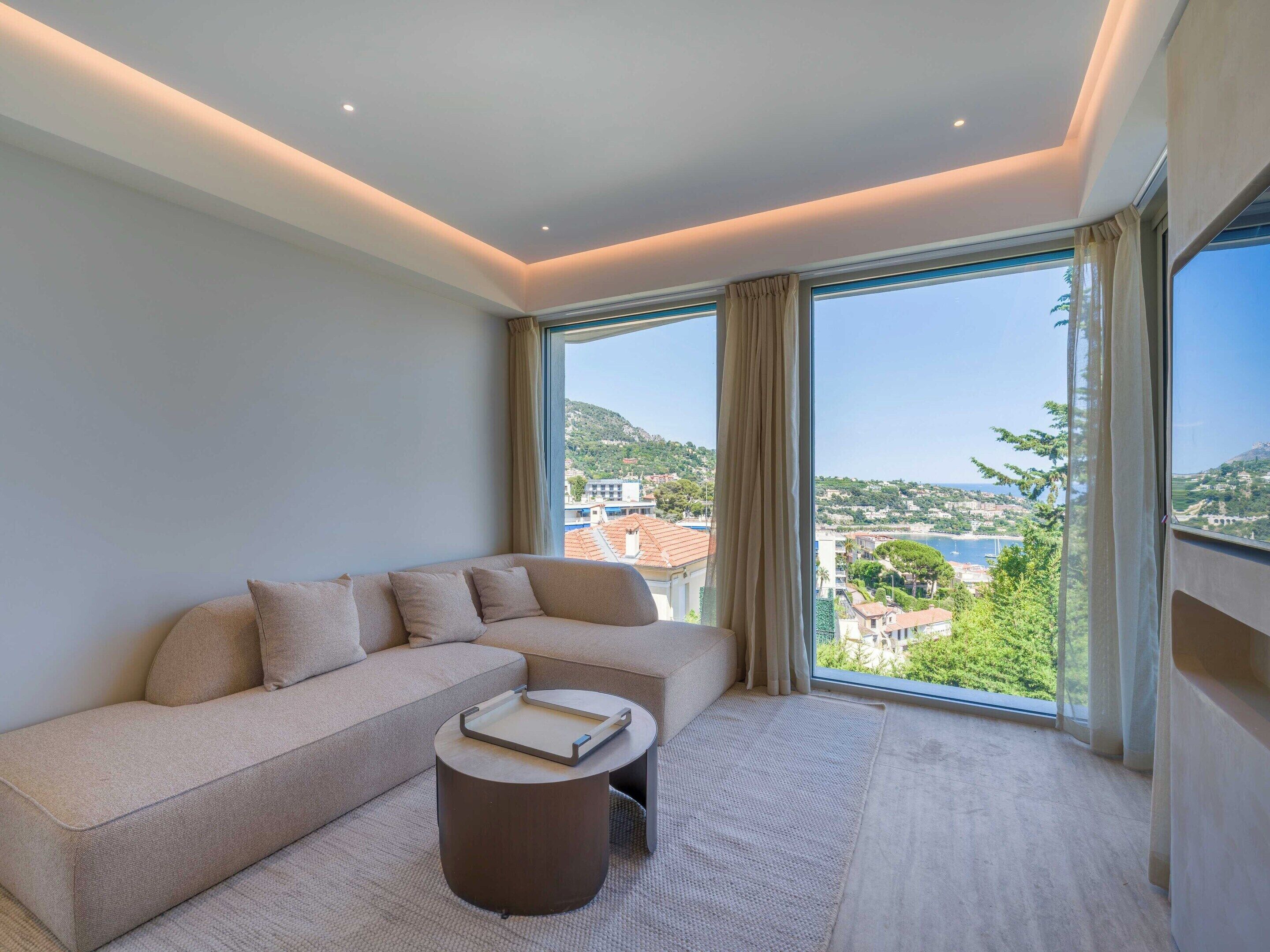 A modern living room with a sofa, a round coffee table, and a large window offering a view of a coastal landscape.