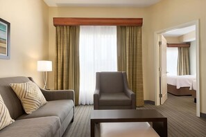 Ein Hotelzimmer mit einer Couch, einem Sessel und einem kleinen Tisch. Eine Tür führt zu einem Schlafzimmer mit einem Bett.