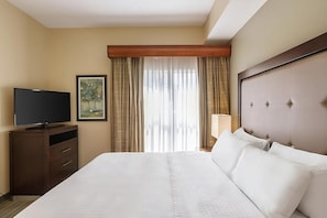 Ein Hotelzimmer mit einem großen Bett, einem Fernseher auf einer Holzkassette, einem Bild an der Wand und einem Vorhangfenster.