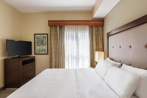 Ein Hotelzimmer mit einem großen Bett, einem Fernseher auf einer Holzkassette, einem Bild an der Wand und einem Vorhangfenster.