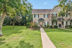A two-story house with a front yard, a pathway leading to the entrance, and a tree with pink flowers. - Salt + Sand - Oceanwalk 3 bedroom pet-friendly (St. Simons Island)