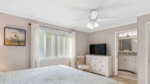 Ein Schlafzimmer mit Bett, Fernseher, Kommode und einem Badezimmer mit Spiegel und Waschbecken.