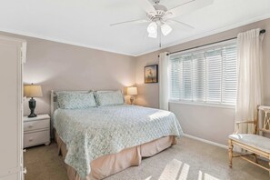 Ein Schlafzimmer mit Bett, Nachttischlampen, einem Nachttisch, einem Sessel, einem Fenster mit Jalousien und einem Deckenventilator.