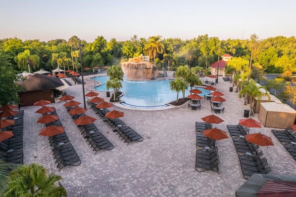 Ein Poolbereich eines Resorts mit mehreren Liegestühlen und Sonnenschirmen, einem zentralen Wasserfall und üppiger, umgebender Bepflanzung.