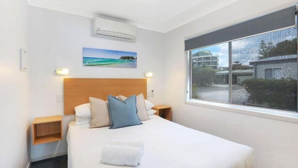 A hotel room with a bed, a window with a view, a painting on the wall, and a small wooden shelf. - Reflections Holiday Park Forster Beach (Forster)