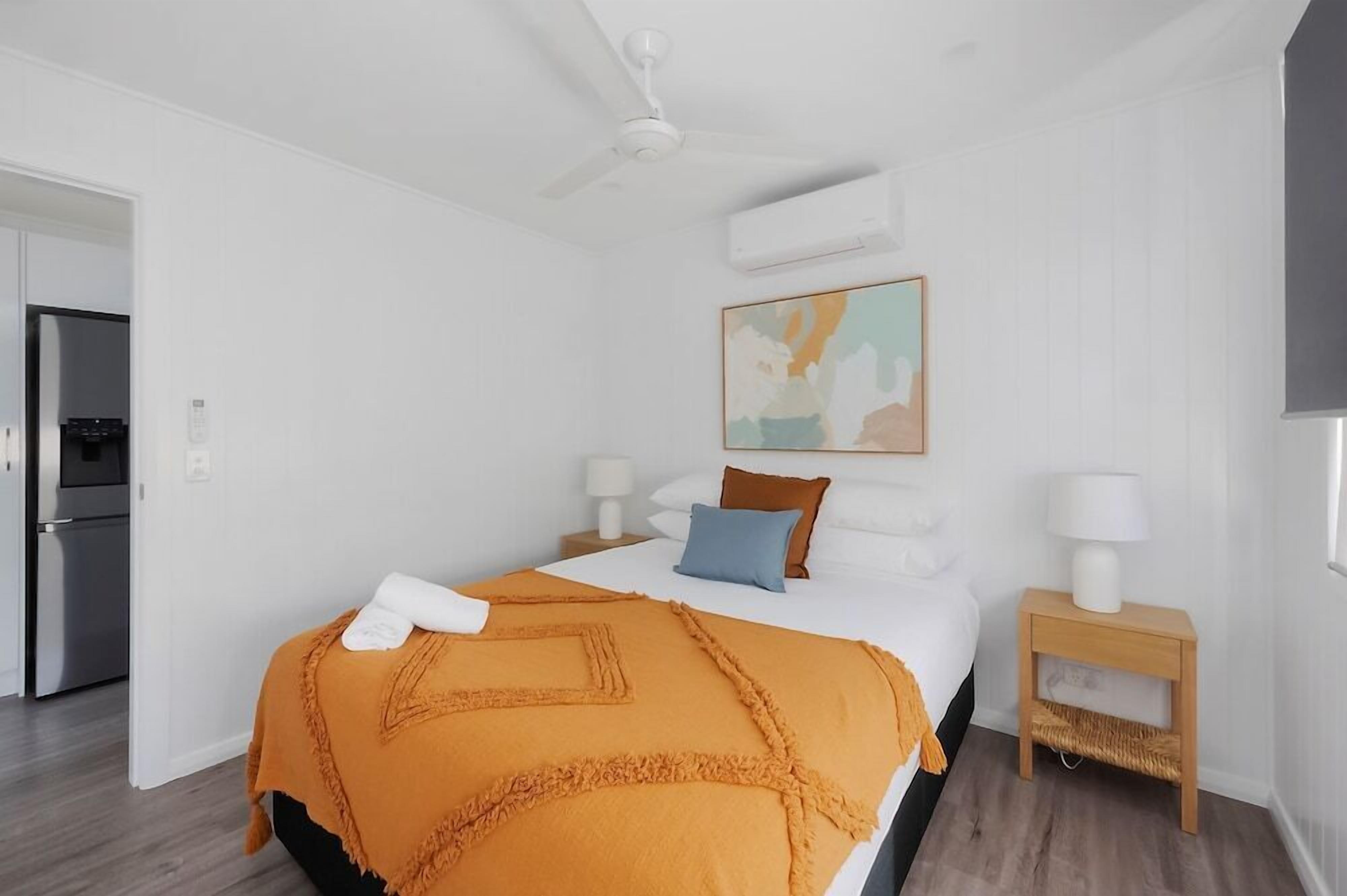 Une chambre d’hôtel avec un lit, un ventilateur de plafond, un appareil de climatisation, un tableau au mur et une petite table de chevet en bois avec une lampe.