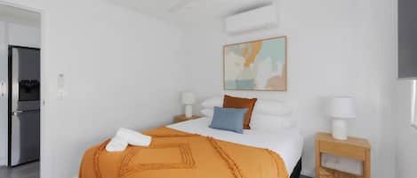 A hotel room with a bed, a ceiling fan, an air conditioning unit, a painting on the wall, and a small wooden nightstand with a lamp.
