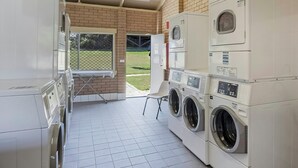 A laundry room with stacked washers and dryers, a table with a chair, and a view of a grassy yard through an open door. - Reflections Holiday Park Forster Beach (Forster)
