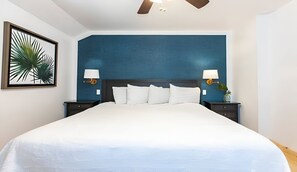 Ein großes Bett mit weißer Bettwäsche, zwei Nachttische mit Lampen, ein Deckenventilator und eine gerahmte Pflanze an der Wand.