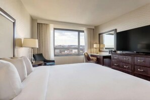 Ein Hotelzimmer mit einem großen Bett, einem Schreibtisch, einem Sessel, einem Fernseher und einem Fenster mit Blick auf die Stadt.