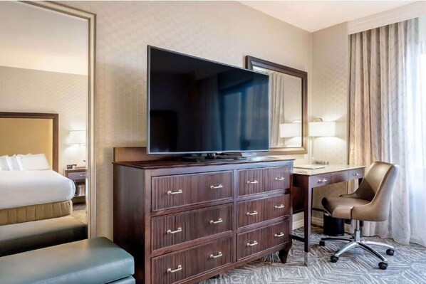 Habitació d'hotel amb un llit, un televisor de pantalla plana gran sobre una petita estanteria de fusta, una taula de treball amb una cadira, i una finestra amb cortines.