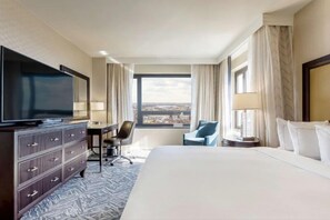 Ein Hotelzimmer mit einem großen Bett, einem Fernseher, einem Schreibtisch mit Stuhl und Blick auf die Stadt durch das Fenster.