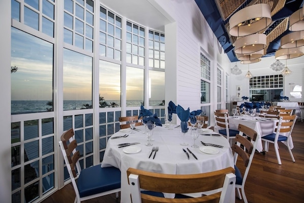 Une salle à manger avec de grandes fenêtres donnant sur la mer, des nappes blanches et des serviettes bleues.