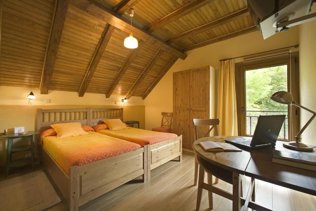 Una habitación con dos camas de madera, un escritorio con una computadora portátil, una silla y un ventanal con cortinas.