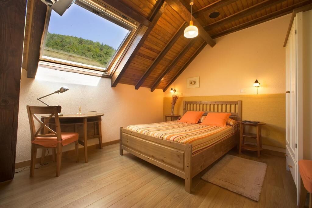 A bedroom with a sloped ceiling, a bed with striped bedding, a desk with a chair, and a window with a view of greenery.