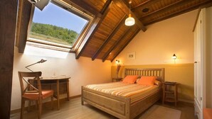 Ein Schlafzimmer mit schrägen Wänden, einem Bett mit gestreifter Bettwäsche, einem Schreibtisch mit Stuhl und einem Fenster mit Blick auf Grünflächen.