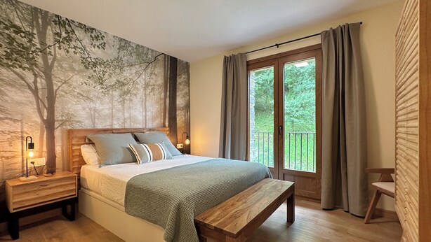 Ein Schlafzimmer mit einem großen Bett, einem Holz-Kopfteil, einer Bank, einem Nachttisch und Blick ins Grüne durch ein Fenster.