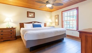 Ein Schlafzimmer mit Deckenventilator aus Holz, einem großen Fenster mit Aussicht, einem Bett mit weißer Bettwäsche und zwei Nachttischen mit Lampen.