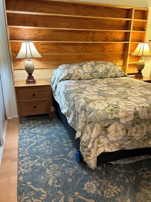 Ein Schlafzimmer mit einem hölzernen Kopfende, einem Bett mit floralem Bettzeug, zwei Nachttischen mit Lampen, einer kleinen Schublade und einem gemusterten Teppich.