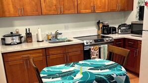 Une cuisine avec des meubles de rangement en bois, une table à manger recouverte d’une nappe à motifs de feuilles, et divers appareils de cuisine.