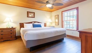 Un dormitorio con ventilador de techo de madera, un ventanal con vistas, una cama con ropa de cama blanca y dos mesitas de noche con lámparas.