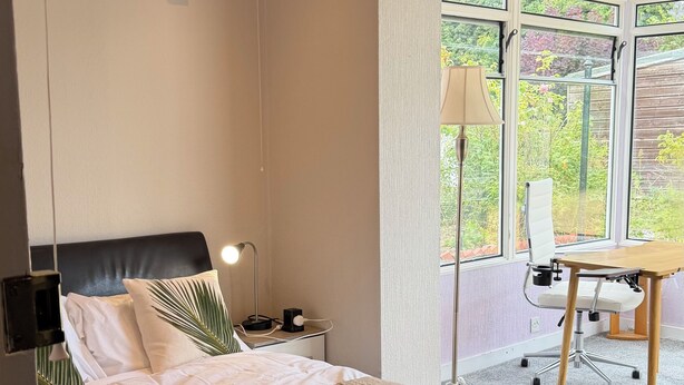 A bedroom with a bed, bedside table, lamp, chair, and a view of greenery through large windows. - Cute cottage for 8, near city centre, parking, gardens. Newly refurbished. (Edinburgh)