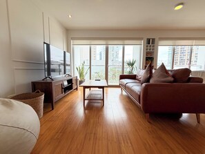Ein modernes Wohnzimmer mit Flachbildfernseher, Holzboden, einem braunen Ledersofa und einem großen Fenster mit Blick auf die Stadt.