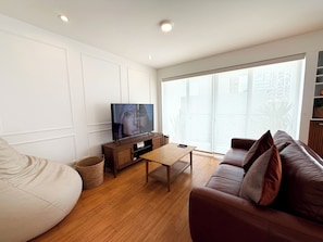 Ein modernes Wohnzimmer mit einem Flachbildfernseher, einem braunen Ledersofa, einem Holztisch und einem großen Fenster mit Blick auf Gebäude.