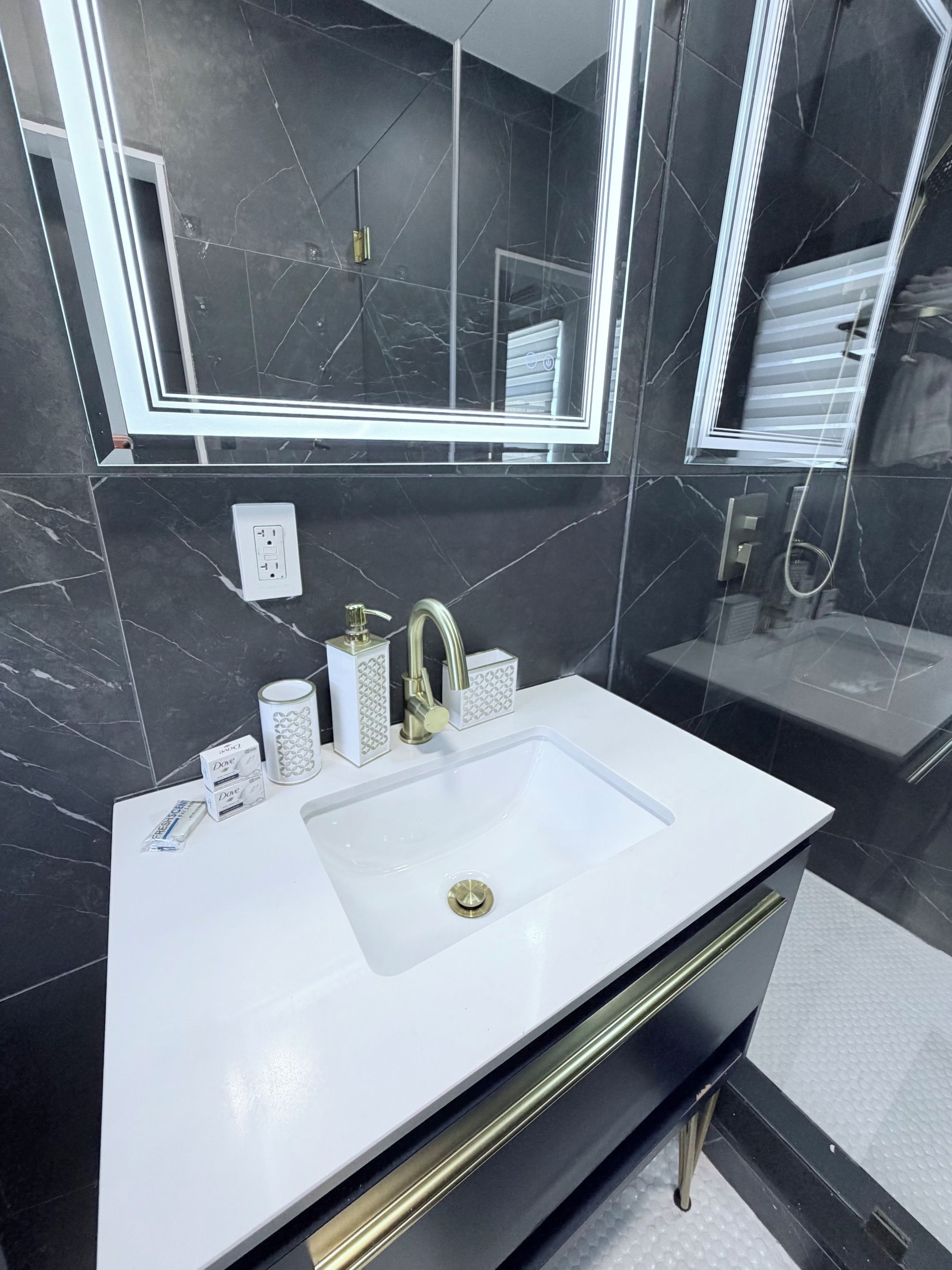 A modern bathroom with a white sink, gold faucet, and a large mirror.