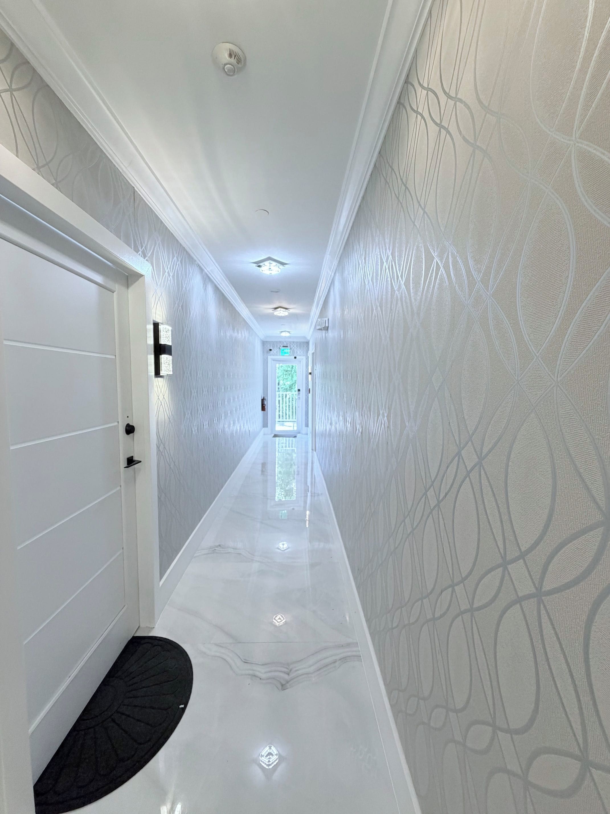 A hallway with a glossy floor, patterned walls, and a door with a black mat.