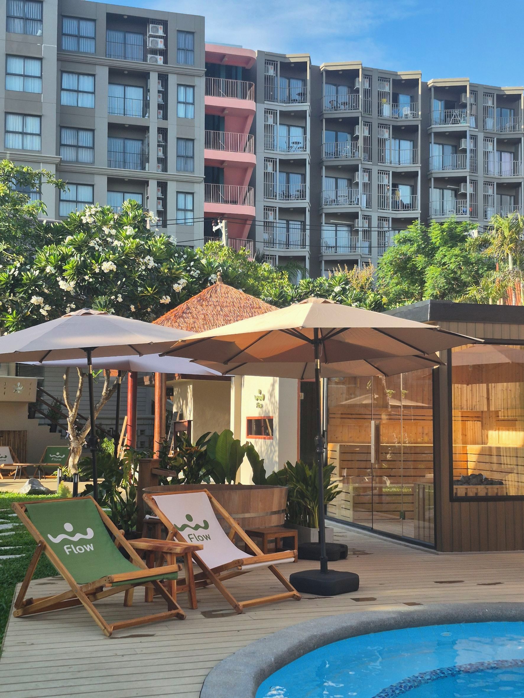 A poolside area with lounge chairs and umbrellas, a wooden deck, and a building with multiple balconies in the background.