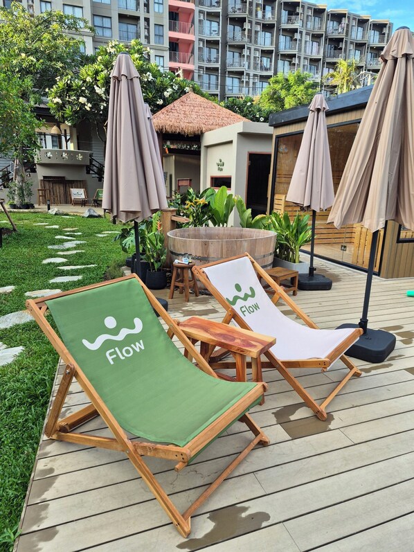 Eine Terrasse mit Liegestühlen und Sonnenschirmen, umgeben von Grünpflanzen und einem Gebäude im Hintergrund.