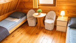 Ein kleines Zimmer mit zwei Einzelbetten, einem kleinen runden Tisch mit Pflanze, zwei gemusterten Stühlen, einem Nachttisch mit Lampe und Holzwänden.