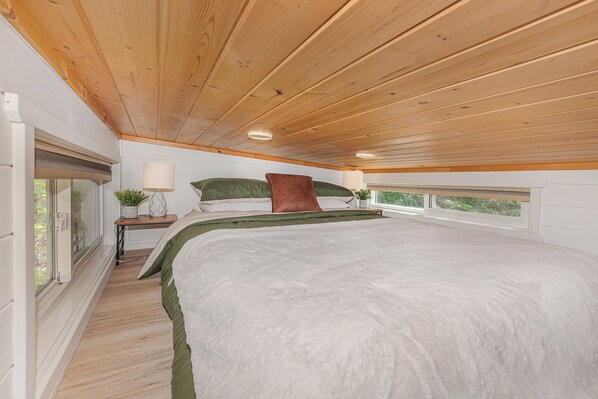 A bedroom with a large bed, a nightstand, a lamp, and a window with a view. - Boutique Tiny Home Escape | Private Hot Tub , Pizza Oven & Gazebo (Swansea Point)