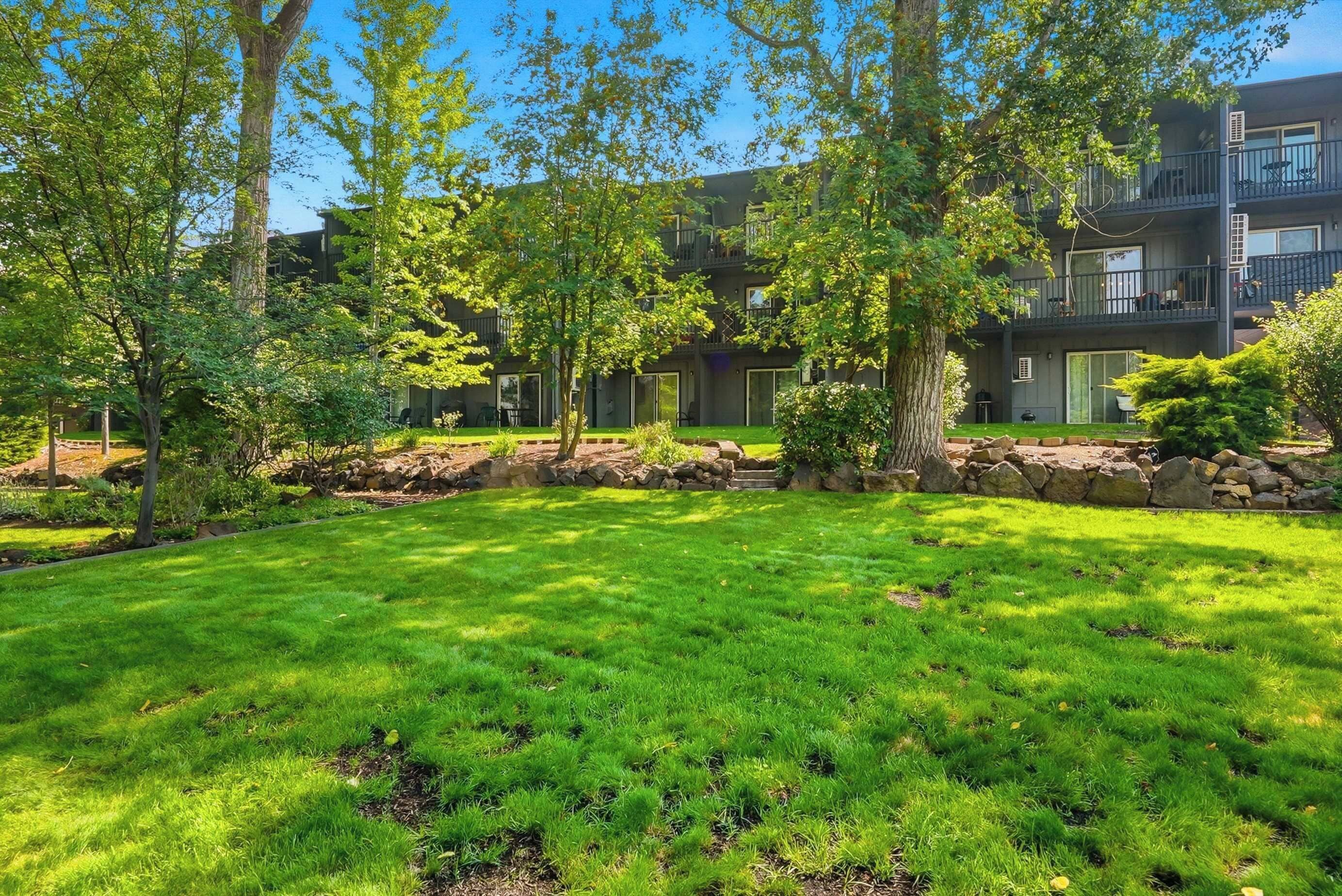 A well-maintained grassy lawn with trees and a stone wall in the foreground, leading to a modern building with balconies and large windows.