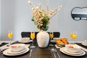 Ein Esstisch ist mit Tellern, Schüsseln, Besteck, Gläsern Orangensaft und einer Blumenvase gedeckt.