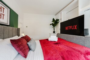 Ein modernes Hotelzimmer mit einem Bett, einer roten Decke, einem grauen Sofa, einer Pflanze und einem an der Wand montierten Fernseher, der 'NETFLIX' anzeigt.