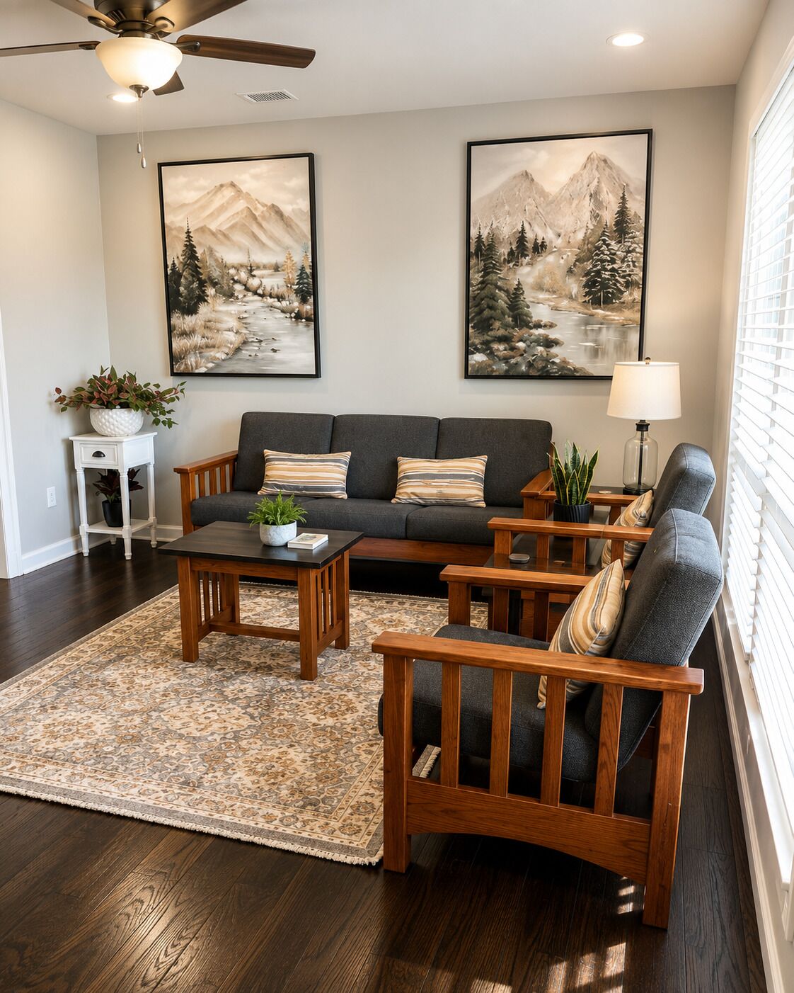 A living room with a dark wood floor, a grey sofa, a wooden coffee table, and two armchairs with cushions. There are two paintings on the wall, a ceiling fan, and a window with blinds.